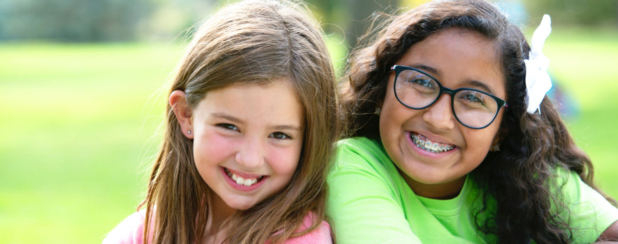 Two preteen girls smile as they sit back to back on a picnic bench.