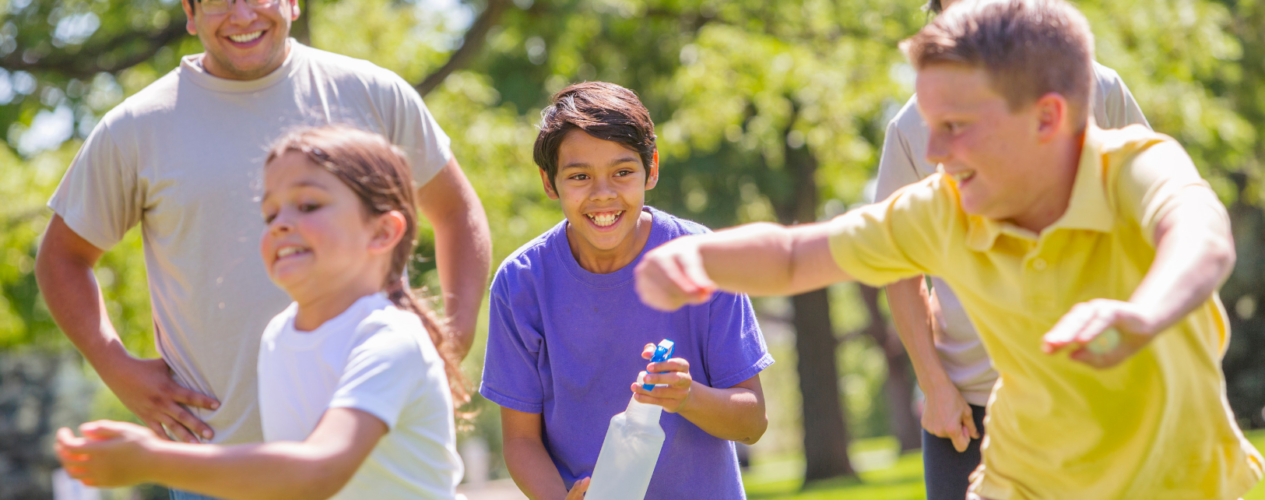Three elementary-aged children playing tag with a spray bottle. An adult volunteer oversees their game.