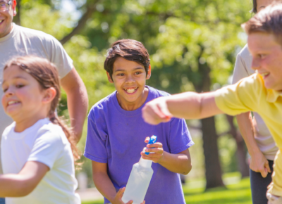 Three elementary-aged children playing tag with a spray bottle. An adult volunteer oversees their game.