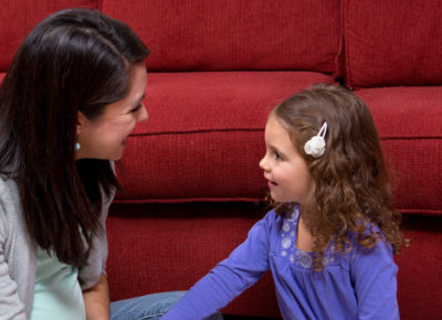 A mom and her preschool daughter sit on the ground in front of a couch, talking.