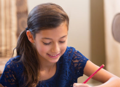 A preteen girl sits at a coffee table writing a letter.