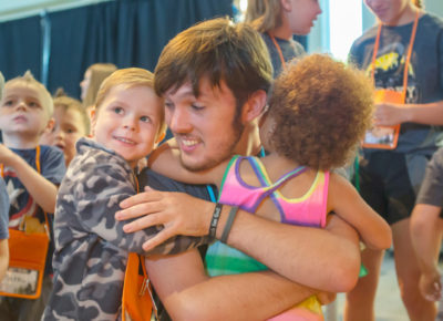 A college-aged volunteer hugs a group of preschool children.