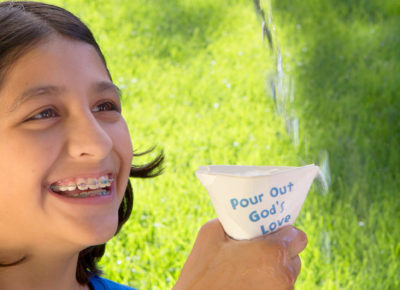An elementary girl is holding a cup as water is poured into it. It is overflowing.