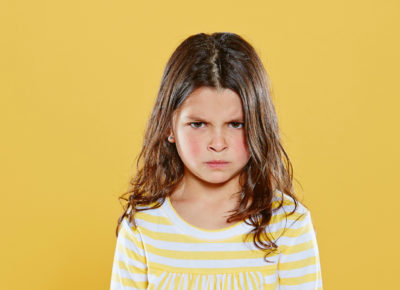 A girl wearing a yellow striped shirt is standing in front of a bright yellow background. She looks like she is one of those children with negative attitudes.