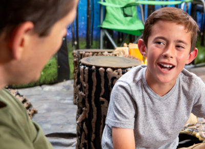 A preteen boy smirks at his small group leader as they talk in front of a indoor camping scene.