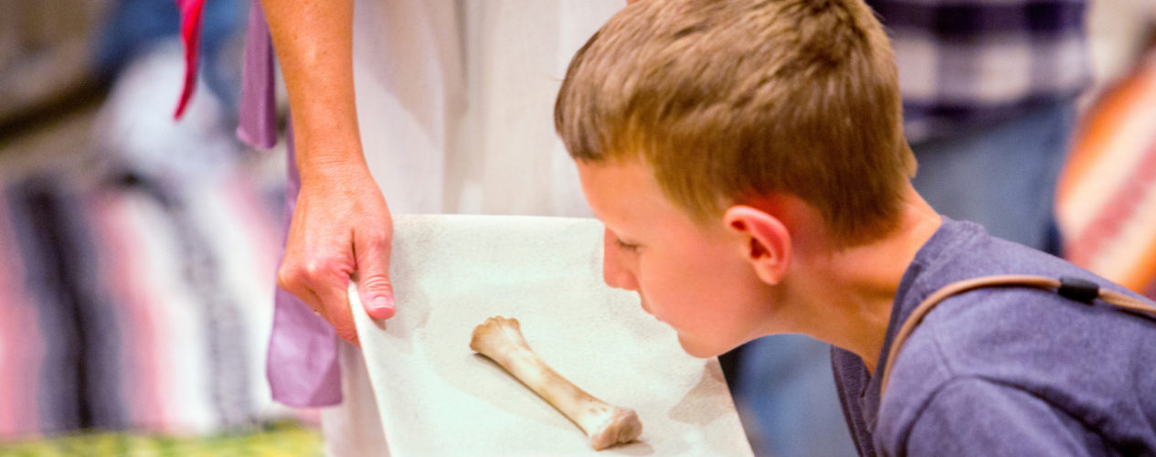 An older elementary boy sits on the ground at a Seder dinner. He is looking at a lamb bone.