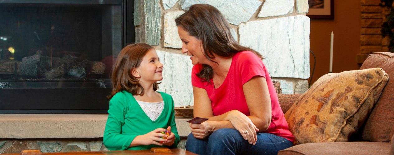 A mom sits in front of a fireplace with her elementary-aged daughter. They are talking and laughing as they get ready to use the encouragement web with the rest of their family.