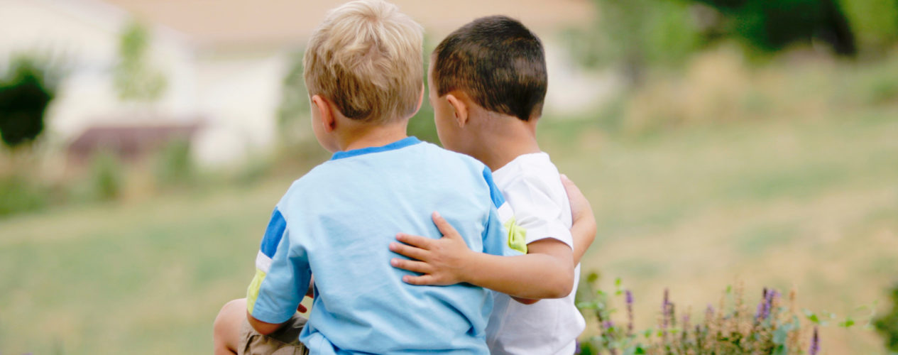 Two kindergarten boys sit on a grassy hill with their arms linked around one another.