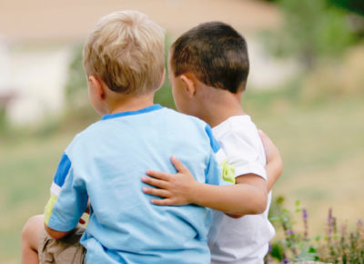Two kindergarten boys sit on a grassy hill with their arms linked around one another.