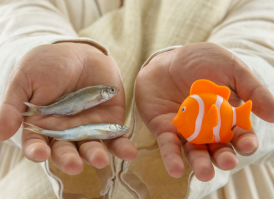 A man in Biblical clothes holding to minnows in one hand and a plastic goldfish in the other.