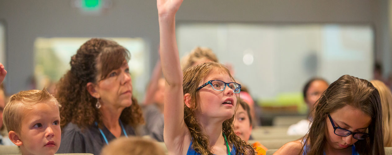 An elementary-aged girl raises her hand during the children's message about help.