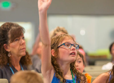 An elementary-aged girl raises her hand during the children's message about help.