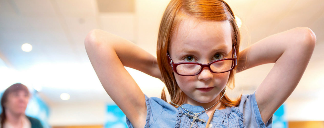 Little girl with glasses is putting both her hands behind her head as she looks over her glasses and down at the camera.