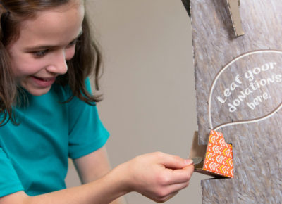 A girl puts a donation envelope in a tree that says "Leaf your donation here."