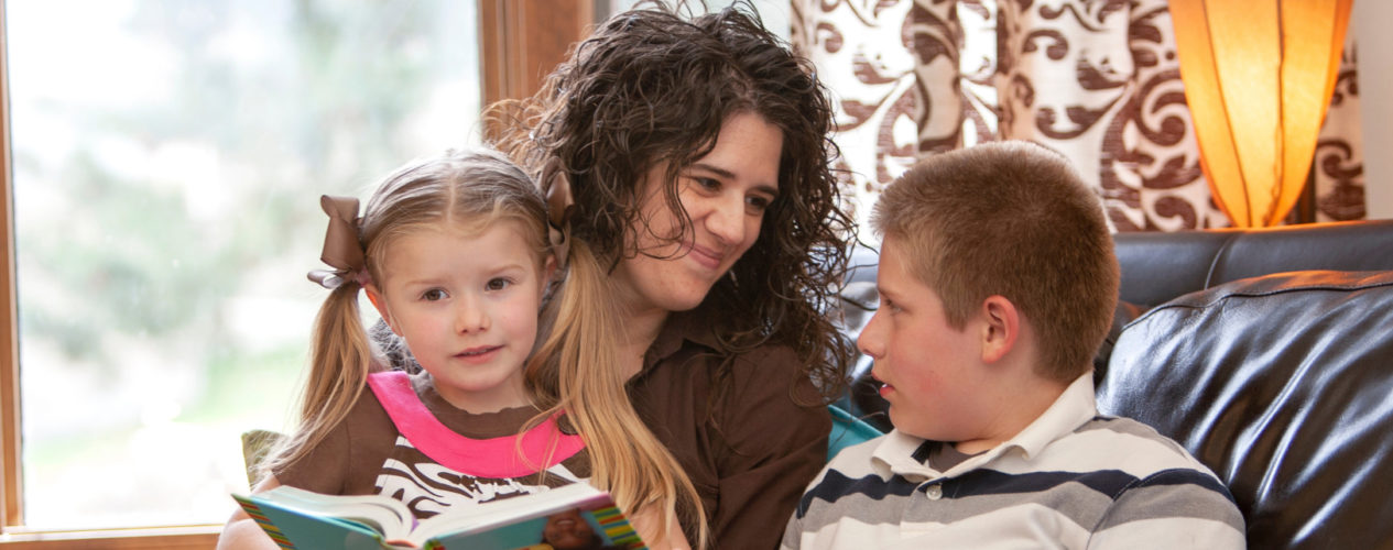 A mom reads the Bible with her two children, a preschool girl who is sitting on her lap and a preteen boy who is sitting next to her.