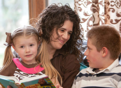 A mom reads the Bible with her two children, a preschool girl who is sitting on her lap and a preteen boy who is sitting next to her.