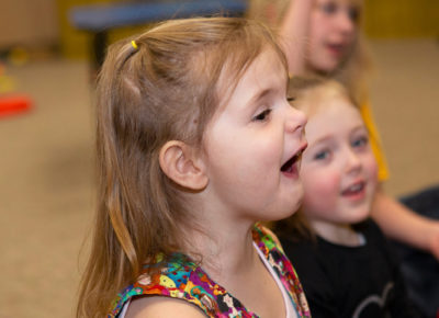 Elementary-aged girl sits among her classmates in children's ministry. She has her mouth wide open in disbelief.