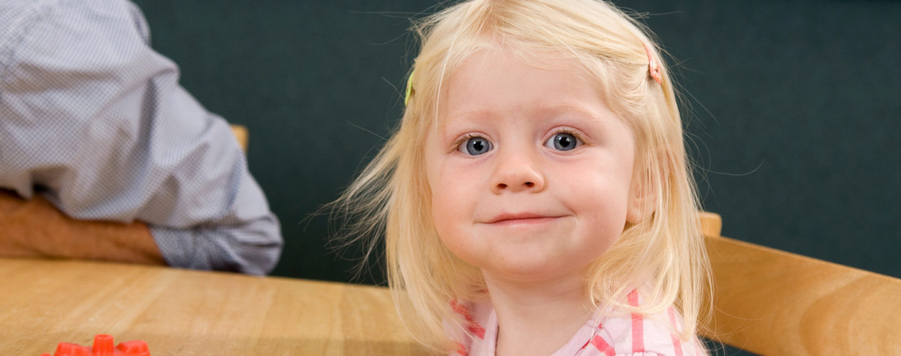 Preschool girl smiles at the camera as she sits at a table waiting for her craft.