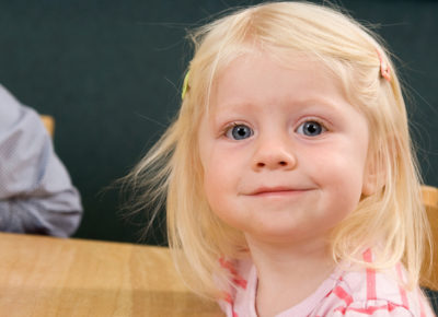 Preschool girl smiles at the camera as she sits at a table waiting for her craft.