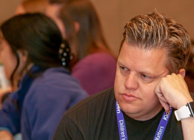 A male volunteer sits at a table. He's rest his check on his hand and has a pouty expression on his face.