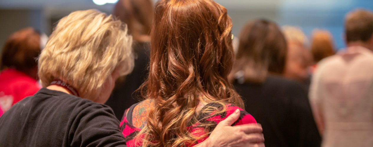 Two women in a worship service. One woman has her arm around another woman's shoulder. They are praying against burnout.