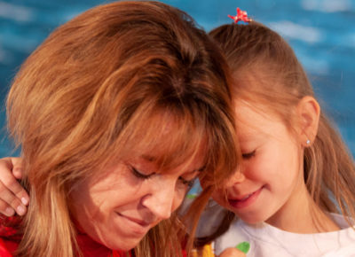 A mom and her preschool daughter are praying together with their heads leaning in towards one another. The daughter has her arm around her mom's neck.