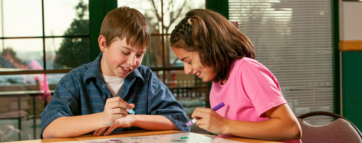 A preteen boy and girl are sitting at a table working a project. The boy is talking and the girl is smiling.