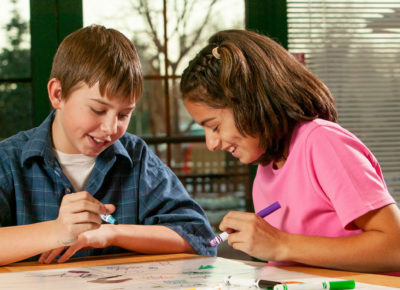 A preteen boy and girl are sitting at a table working a project. The boy is talking and the girl is smiling.