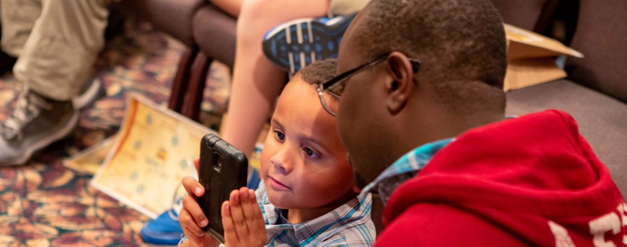 A dad and son sitting on the ground together in a church sanctuary. They are looking at something on the dad's phone.
