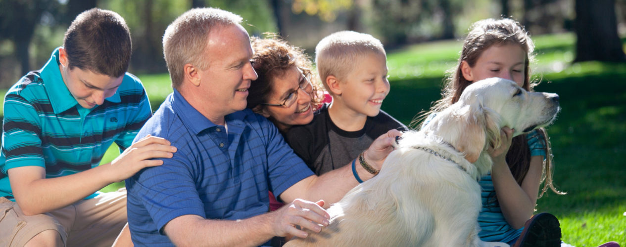 A family with a dad, mom, and three kids sitting outside on the grass. They are petting a yellow lab.
