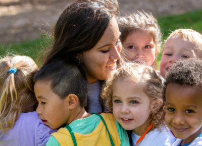 A woman volunteer sits on the gras as six preschoolers surround her in a group hug.