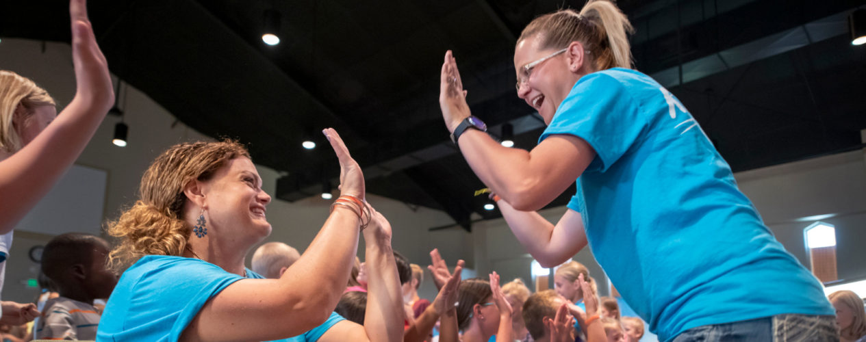 Two adult women high five in the main session of a VBS. There are kids all around, very joyful!
