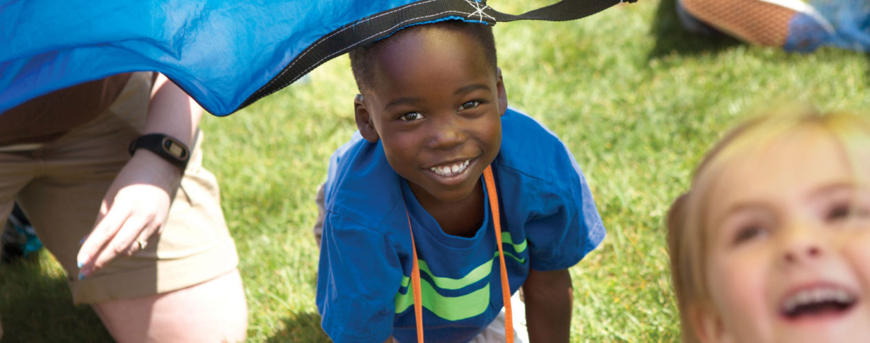 An elementary-aged VBS visitor is crawling out from under a colorful parachute. He's smiling!