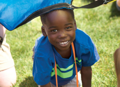 An elementary-aged VBS visitor is crawling out from under a colorful parachute. He's smiling!