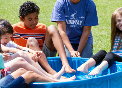 A group of kids are sitting outside on the grass with their feet in a kiddie pool. They're trying to grab ice cubes with their toes!