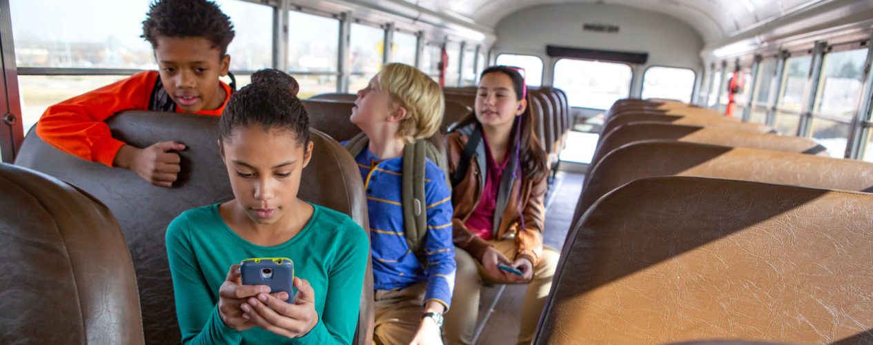 A preteen girl sitting on the bus staring with concern at her phone. A boy is looking over her shoulder and there are a few other kids on the bus.