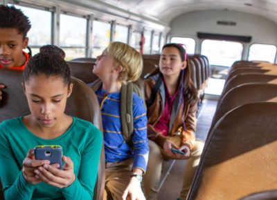 A preteen girl sitting on the bus staring with concern at her phone. A boy is looking over her shoulder and there are a few other kids on the bus.