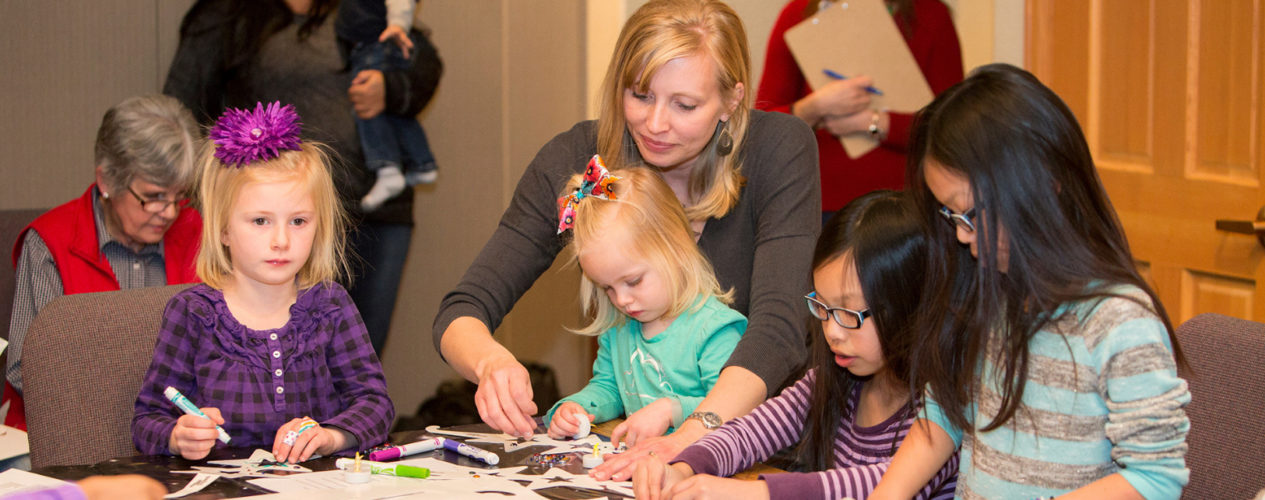 A woman volunteer helps a table of girls ranging from a toddler to two 3rd graders participate in a Christmas craft.