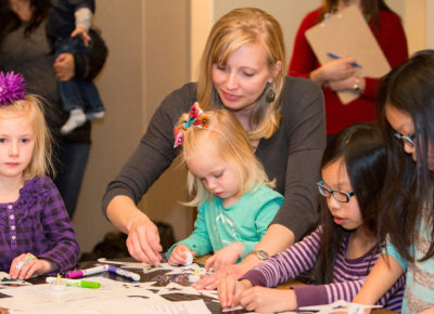 A woman volunteer helps a table of girls ranging from a toddler to two 3rd graders participate in a Christmas craft.