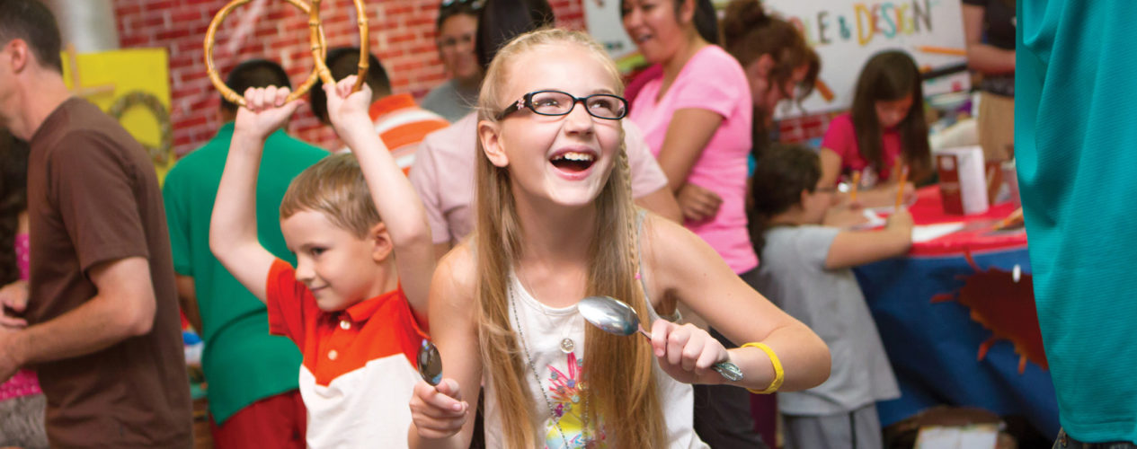 An older elementary aged girl is smiling as she and her brother sing and dance at a fall fest. She is holding a spoon in her hand that she was using as a musical instrument.