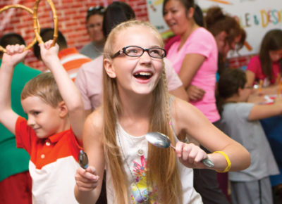 An older elementary aged girl is smiling as she and her brother sing and dance at a fall fest. She is holding a spoon in her hand that she was using as a musical instrument.