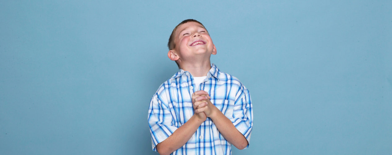 Elementary aged boy in a blue checkered shirt. He's praying as he smiles up towards God.