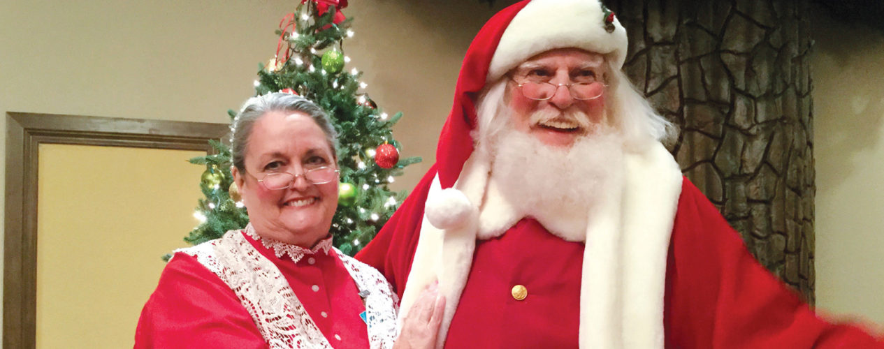 A man and woman dressed as Santa and Ms. Clause. They are standing joyfully in front of a Christmas tree.