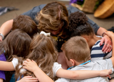 A group of children sitting on the ground in a circle with their small group leader. They have their arms around one another and are praying.