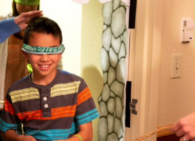 A preteen boy in a polo with large, colorful stripes is blind-folded as he walks through a door.