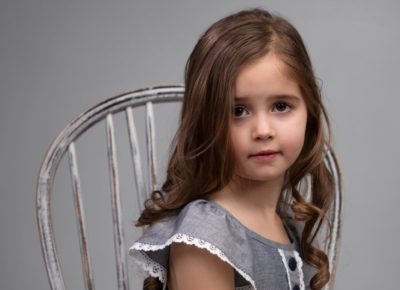 A sad looking elementary girl sitting on a chair in front of a gray background.