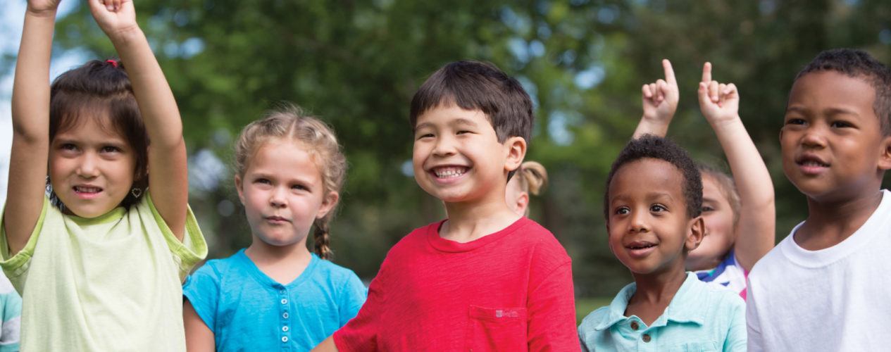 A group of kids outside smiling. The kids are all different heights and are diverse in ethnicity.