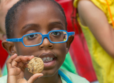 A boy wearing blue glasses smiles as he holds up a cookie bite.