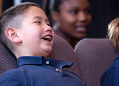 An elementary-aged boy in a navy polo is laughing as he sits in a chair. Other kids are around him and listening as well to the candy cane story being told.
