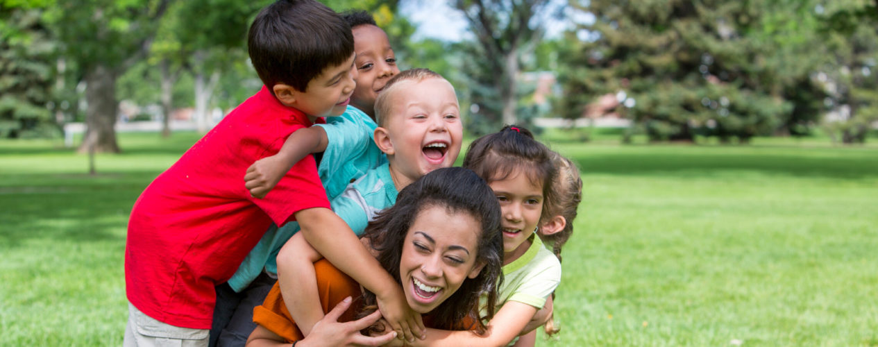 A group of four children laughing as they climb on their children's ministry volunteer.